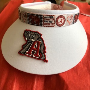 May include: A white visor with a decorative band featuring the text "Roll Tide" and "Love God, Sweet Tea and the Crimson Tide." An embroidered elephant and the letter "A" are on the front.