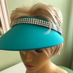May include: A teal visor with a wide brim and a row of rhinestones around the band.