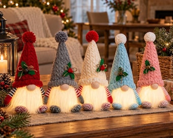 Puede incluir: Cinco figuras de gnomos de peluche con barbas blancas iluminadas, cada una con un gorro alto de punto trenzado en rojo, gris, crema, azul claro o rosa. Cada gorro tiene un pompón y está decorado con hojas de acebo verdes y bayas.