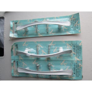 May include: Three packages of white and silver cabinet handles. Each package contains a white handle with silver accents, screws, and the text "A-102-C1 CERAMOID WHITE-GOLDEN ACCENT Priced on back". The handles are on a teal background.