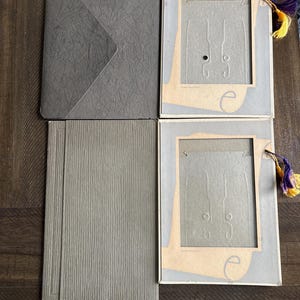 May include: An overhead shot shows two gray photo frames with decorative tassels, a gray envelope, and a gray textured notebook. The frames have a light gray border and a beige interior. The tassels are yellow, purple, and gold.