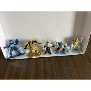 May include: A collection of five colorful action figures, each with unique designs and poses. The figures are in a white box, set against a wooden surface. The figures are in shades of blue, gold, and yellow.
