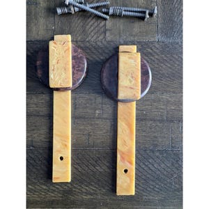 May include: Two vintage cabinet pulls with a light brown, marbled finish and a dark brown circular base. Each pull has a rectangular center piece and a long, rectangular handle. Several screws are visible at the top of the image.