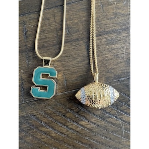 May include: Two gold-colored necklaces. One features a green and gold letter "S" pendant. The other has a gold football pendant with silver and gold accents. Both necklaces are on a wooden surface.