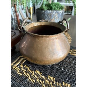 May include: A vintage copper cauldron with two arched handles. The pot has a rounded body and a wide opening. The copper has a warm, aged patina. The cauldron sits on a black and gold woven mat, suggesting a rustic or antique theme.