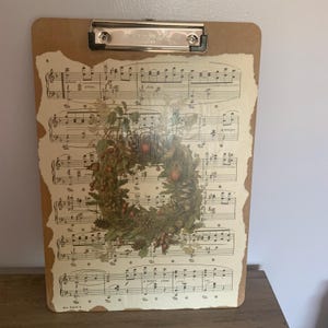 May include: A brown clipboard with a vintage music sheet design featuring a wreath of green leaves, red berries, and pine cones. The sheet music has the words "a tempo" and "dim." printed on it.