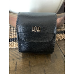 May include: A black leather case with the word "HOYA" in silver lettering. The case is rectangular with a flap closure and stitched edges. The case is sitting on a wooden surface.