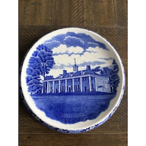 Puede incluir: Un plato decorativo redondo azul y blanco con una ilustración detallada de una casa señorial, probablemente un edificio histórico. El diseño del plato incluye un cielo azul con nubes y árboles, con las palabras "MOUNT VERNON" en la parte inferior.