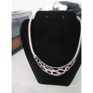 May include: A silver-toned necklace with a unique openwork design. The necklace features a flexible, snake-like chain that curves around the neck. The focal point is a decorative, geometric pendant with an intricate, open pattern. The necklace is displayed on a black velvet bust.
