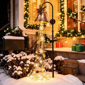 May include: A copper-coloured metal bell hanging from a black shepherd's hook, adorned with red berries and green foliage. Cascading from the bell are warm white LED string lights featuring snowflake shapes and small gold spherical ornaments.