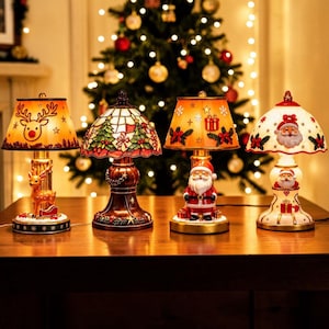 May include: Four decorative table lamps with holiday themes. The first features a reindeer, the second a stained-glass style tree pattern, the third a Santa Claus figure, and the fourth a white base with a Santa face and gift motifs.