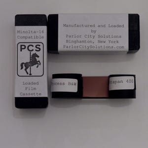 May include: A Minolta-16 compatible film cassette with a white label featuring a carousel horse logo and the text PCS. A second black cassette box is labeled with manufacturer details for Parlor City Solutions. A strip of film is also visible.