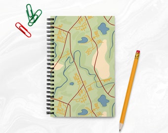 May include: A spiral bound notebook with a green and brown map design. The map features a winding river, roads, and small lakes. The notebook is closed and has a white background.