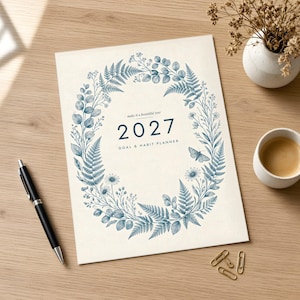 May include: A 2027 goal and habit planner with a cream-colored cover featuring a blue botanical wreath illustration of ferns, leaves, and a moth. The text reads: make it a beautiful year 2027 GOAL & HABIT PLANNER. The planner is rectangular.