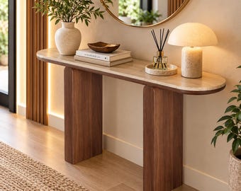 May include: A console table with a light-colored, oval-shaped top and two thick, dark wood legs. The table holds a textured vase with greenery, a stack of books, a small wooden bowl, a reed diffuser, and a mushroom-shaped table lamp.