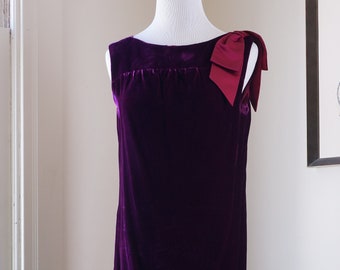 May include: A purple velvet dress with a burgundy bow on the shoulder. The dress is on a black stand.