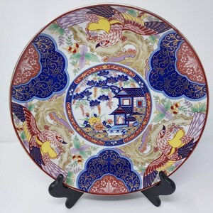 May include: A decorative porcelain plate featuring a central circular medallion with a house and pine tree, surrounded by three phoenix birds, floral motifs, and intricate blue and red patterns with gold accents on a white background.