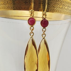 May include: Gold dangle earrings with teardrop-shaped topaz stones and small red ruby accents.
