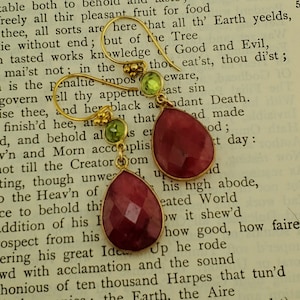 May include: A pair of gold earrings with red gemstone drops and green gemstone accents. The earrings are photographed on a page of a book.