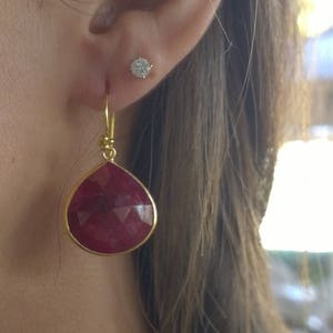 May include: A close-up of a person's ear adorned with two earrings. One is a small, round diamond stud. The other is a gold-framed, teardrop-shaped earring with a faceted, deep red gemstone.