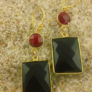 May include: A pair of gold earrings with black and red gemstone accents. The earrings feature a square-shaped black gemstone and a round red gemstone.