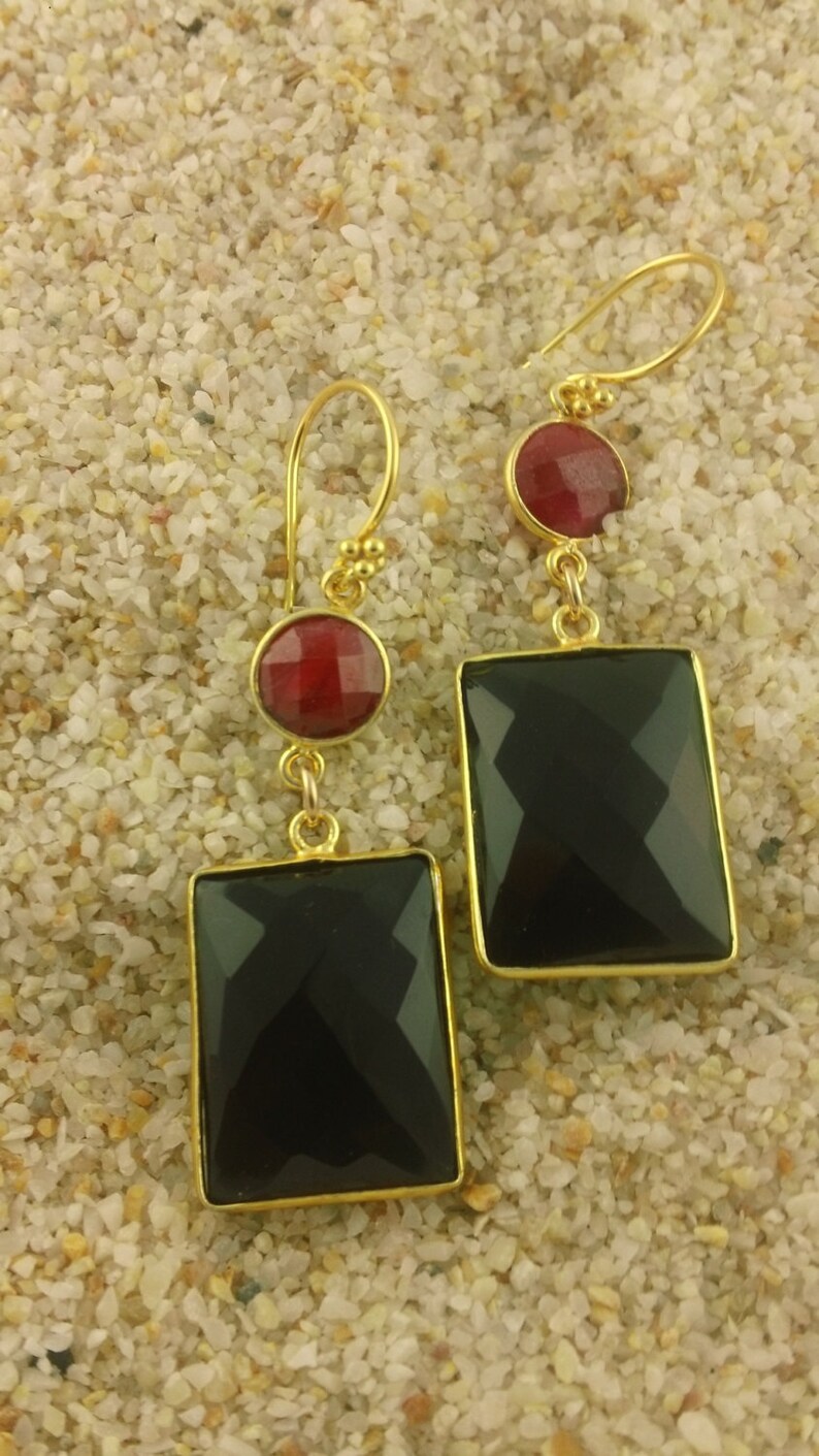 May include: A pair of gold earrings with black and red gemstone accents. The earrings feature a square-shaped black gemstone and a round red gemstone.
