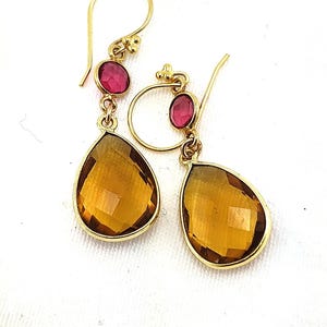 May include: A pair of gold-colored drop earrings. Each earring features a small, oval, pink gemstone at the top, connected to a larger, teardrop-shaped, amber-colored gemstone. The earrings are displayed against a white background.