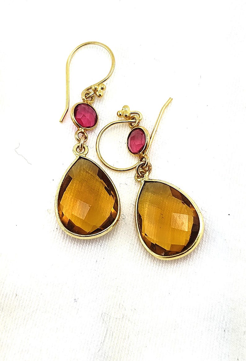May include: A pair of gold-colored drop earrings. Each earring features a small, oval, pink gemstone at the top, connected to a larger, teardrop-shaped, amber-colored gemstone. The earrings are displayed against a white background.