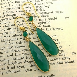 May include: A pair of gold dangle earrings with green gemstone drops. The earrings are on a book with text.