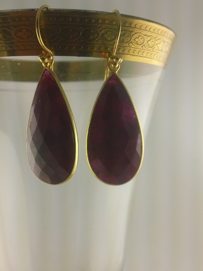 May include: A pair of gold dangle earrings with a teardrop-shaped purple gemstone.