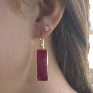 May include: Gold-toned rectangular earrings with a deep red, gem-like center. A small, round, clear stud earring is also visible. The earrings are hanging from a person's ear.