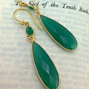May include: A pair of gold earrings with green gemstone drops. The earrings have a teardrop shape and are accented with smaller round green gemstones.