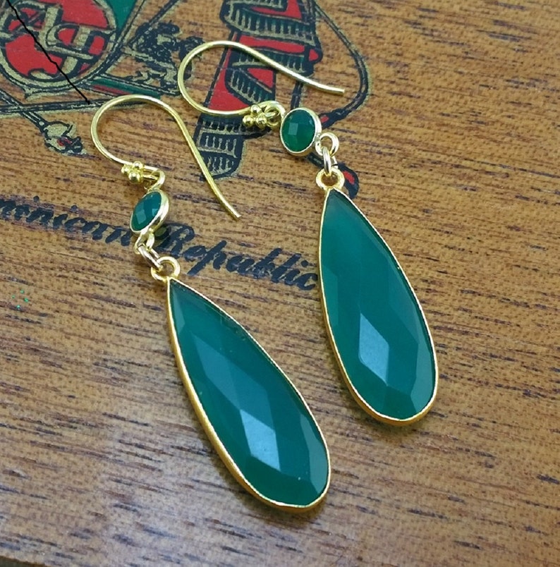 May include: A pair of gold-toned dangle earrings with green gemstone drops. The earrings feature a smaller round green gemstone at the top of each drop.