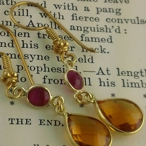 May include: A pair of gold earrings with teardrop-shaped amber stones and round pink stones. The earrings are hanging from a book with the text "BOOK III" and "THE END."