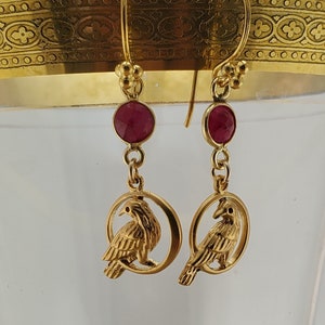 May include: Gold earrings with a red gemstone and a bird design. The bird is perched on a circular branch.