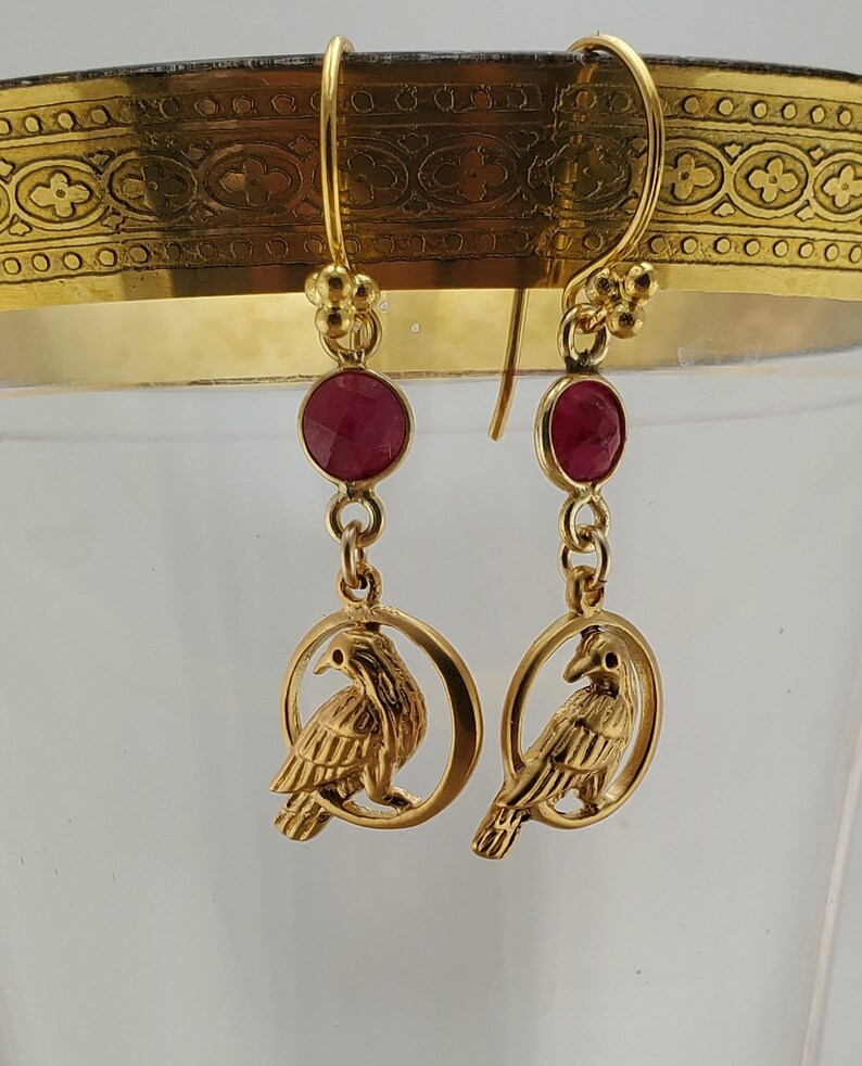 May include: Gold earrings with a red gemstone and a bird design. The bird is perched on a circular branch.