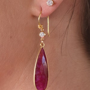 May include: Gold dangle earrings with a teardrop-shaped ruby gemstone and small round diamonds. The earrings have a gold hook closure.