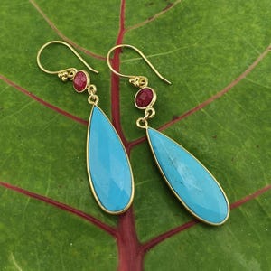 May include: A pair of gold-colored earrings with turquoise teardrop-shaped stones and small red gemstones. The earrings are displayed on a green leaf with red veins.