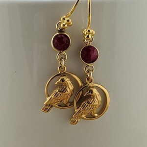 May include: Gold earrings with a red gemstone and a detailed raven design. The raven is perched inside a circle.