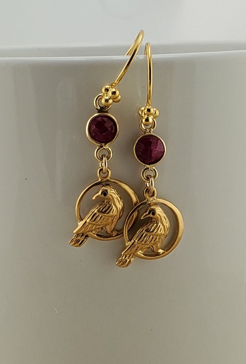 May include: Gold earrings with a red gemstone and a detailed raven design. The raven is perched inside a circle.
