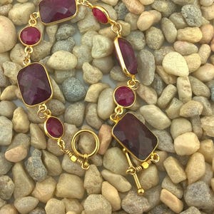 May include: A gold-toned bracelet featuring rectangular and round, deep red gemstones. The bracelet is set against a background of small, smooth, multicolored stones. The clasp is a gold-toned toggle.