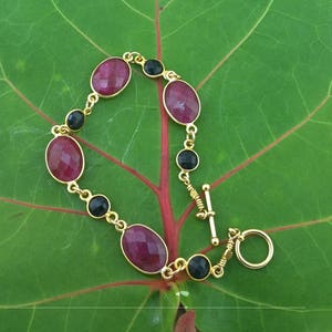 May include: A gold bracelet with alternating oval-shaped red and black gemstones. The bracelet has a toggle clasp.