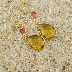 May include: Gold-toned drop earrings with a pink gemstone accent and a larger, yellow-toned, teardrop-shaped gemstone. The earrings are displayed on a sandy surface.
