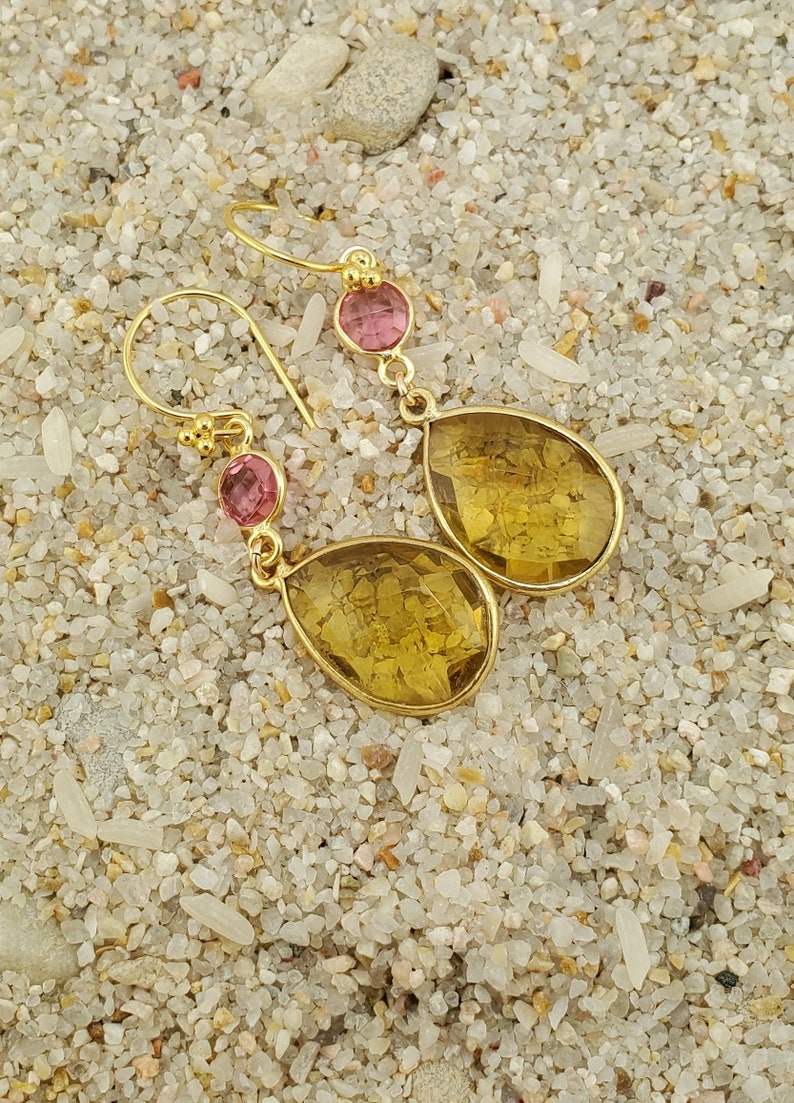 May include: Gold-toned drop earrings with a pink gemstone accent and a larger, yellow-toned, teardrop-shaped gemstone. The earrings are displayed on a sandy surface.