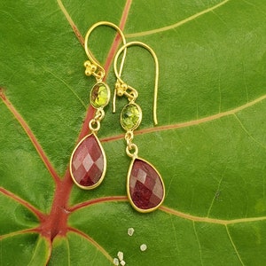 May include: Gold-tone earrings featuring a pear-shaped, faceted, deep red gemstone below a smaller oval, light green gemstone. The earrings have a simple hook design and are displayed on a large green leaf.