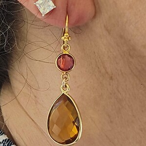 May include: Gold-toned drop earrings with a large, faceted, amber-colored teardrop-shaped stone. A smaller, round, red-toned stone sits above the teardrop. A square, clear stone stud earring is also visible.