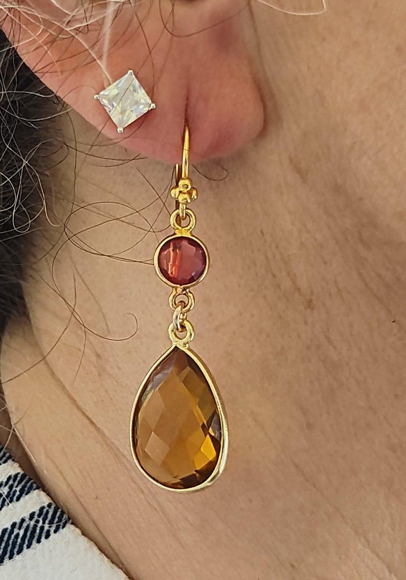 May include: Gold-toned drop earrings with a large, faceted, amber-colored teardrop-shaped stone. A smaller, round, red-toned stone sits above the teardrop. A square, clear stone stud earring is also visible.