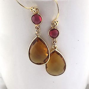 May include: Gold-tone drop earrings with a red gemstone and a large, teardrop-shaped, amber-colored stone. The earrings have a simple hook design and are displayed against a white background. These earrings are a stylish accessory.