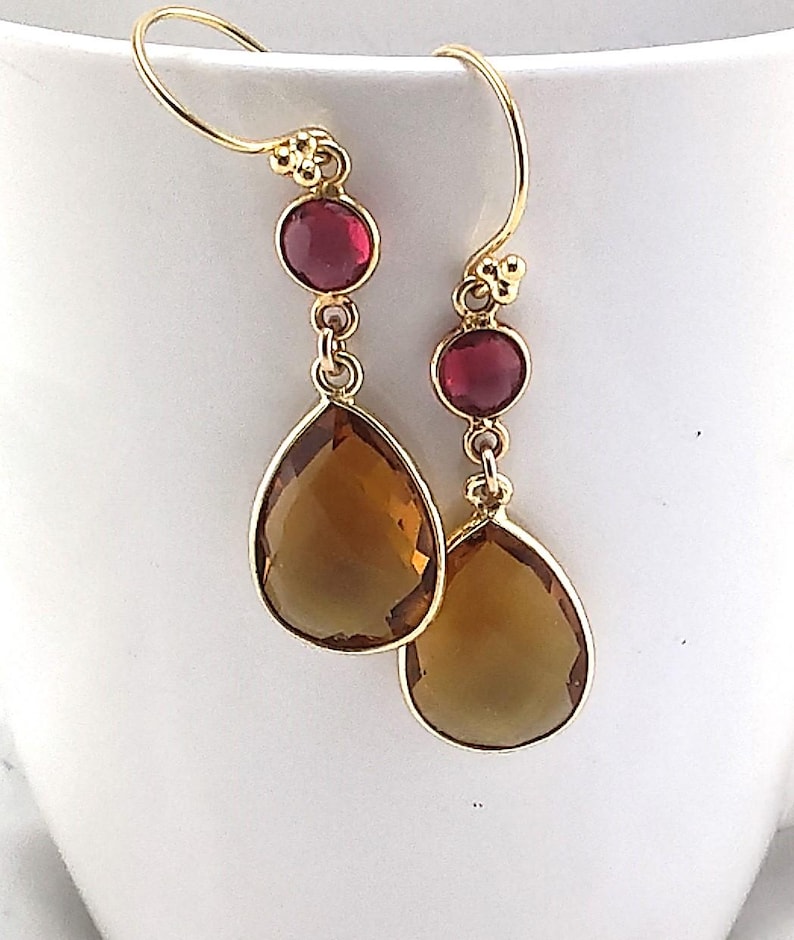 May include: Gold-tone drop earrings with a red gemstone and a large, teardrop-shaped, amber-colored stone. The earrings have a simple hook design and are displayed against a white background. These earrings are a stylish accessory.