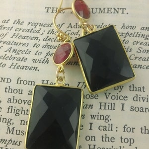 May include: Gold dangle earrings with black square gemstone drops and red gemstone accents. The earrings are photographed on a book with the text "Paradise Lost Book VII."